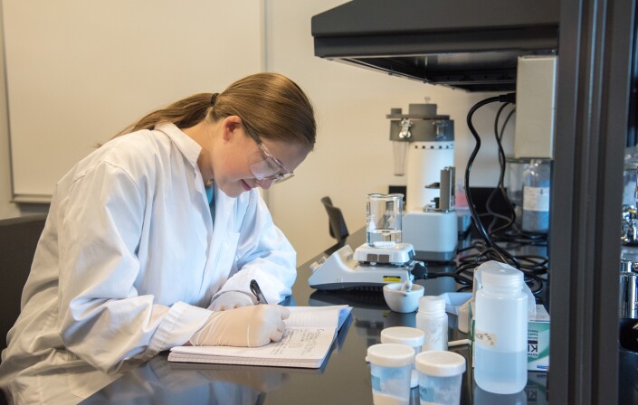 A women wearing a lab coat and goggles, in a lab, writing notes in a notebook.