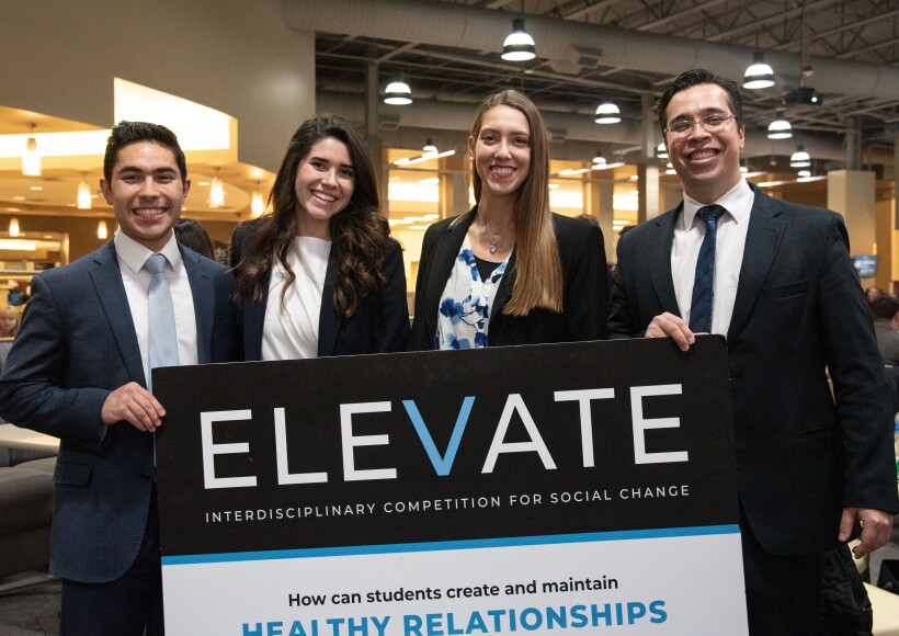 Four BYU-Idaho students in professional attire smile next to an ELEVATE sign during the competition finals.