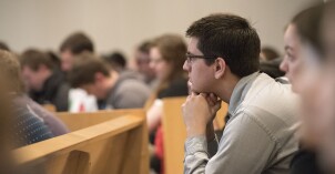 a man leaning forward with his elbow on his knees and his fists under his chin, sitting in a pew, listening to a speaker
