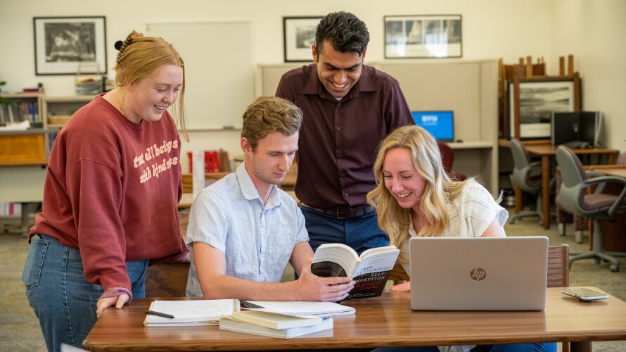 A group of students sitting and standing around a table in the Reading Center looking at a book.
