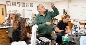 A professor examines something in a classroom with students working with microscopes.