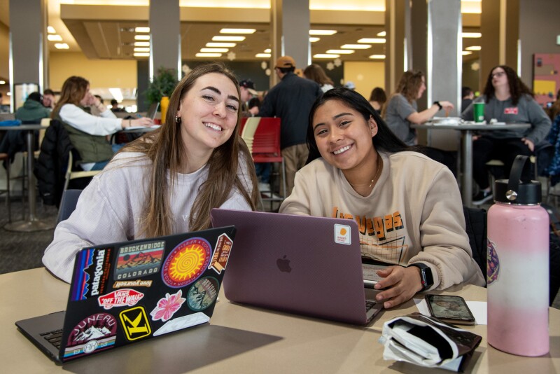 Two students sitting in the Manwaring Center Crossroads, working on their computers and smiling at the camera.