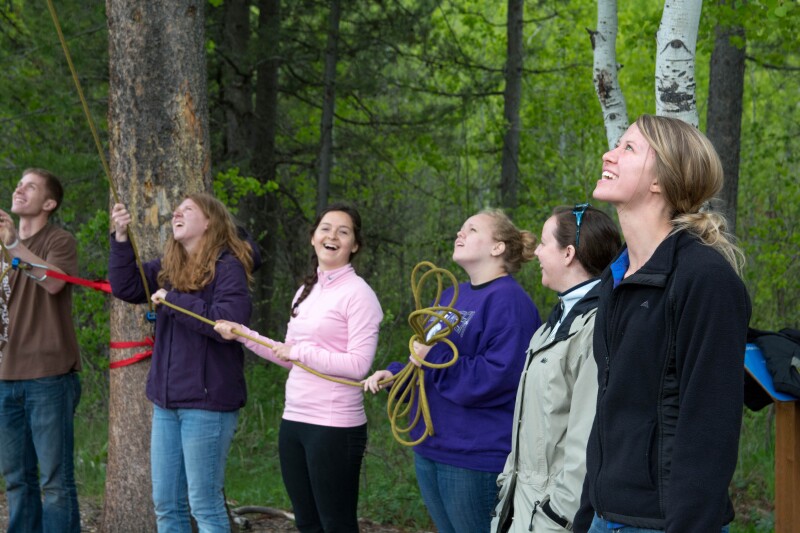 Six BYU-Idaho students look up at a student rock climbing in a forest during a THRIVE activity.