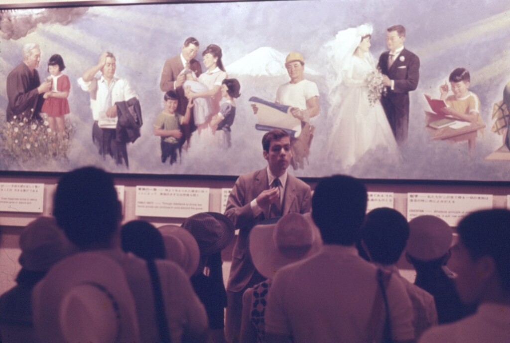 Elder Gessel teaches and stands in front of a painting teaching a crowd.