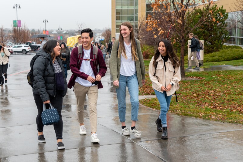 A group of BYU-Idaho students smiling and walking on campus towards the Manwaring Center on a rainy day.