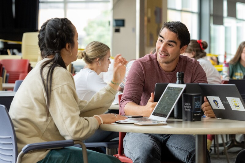 Students sitting at a table in the MC Crossroads, talking to one another.