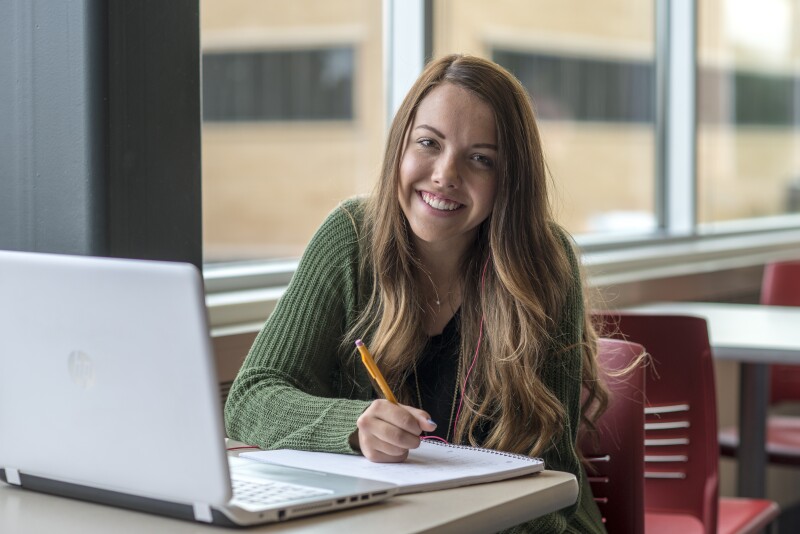 A female student smiles while studying. She is holding a pencil and writing on a pad of paper. An open laptop is on the desk.