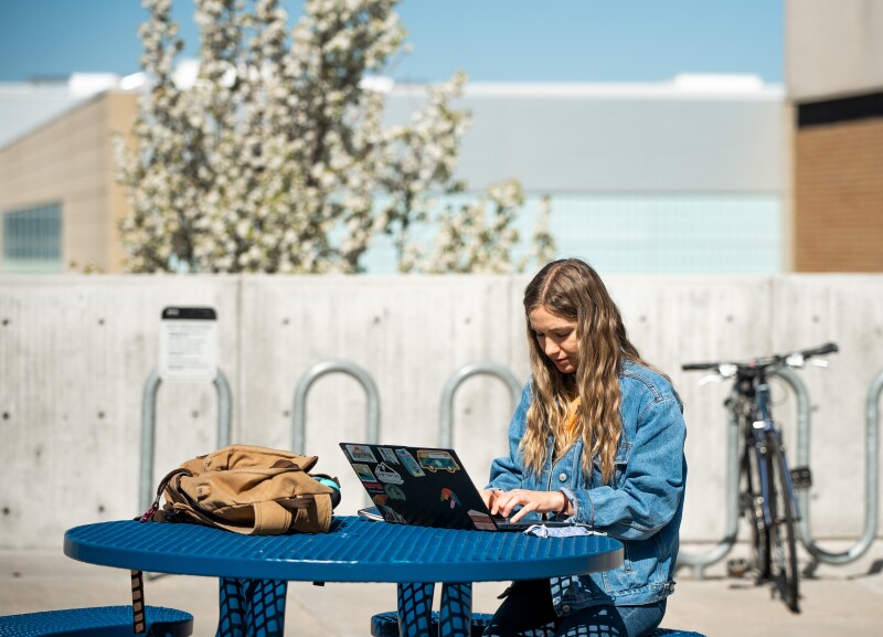 Photo pictures a BYU-Idaho student sitting at a table outside, working on a laptop.