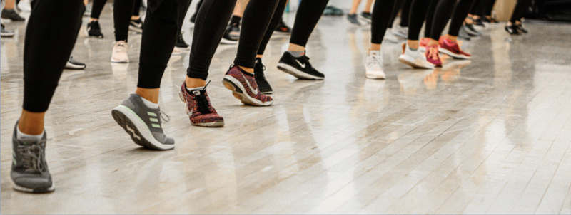 The feet of female students line across a wood-planked floor as they exercise in tennis shoes.