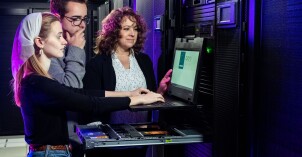 A BYU-Idaho student works on a cybersecurity program while another BYU-Idaho student and a class instructor observe.