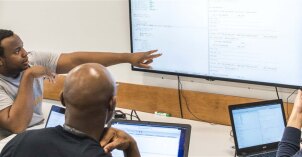 A group, sitting around a table with computers, looking at some information on a bigger tv screen in front of them all