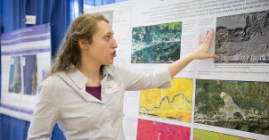 A female student points to diagrams on a wall as she shares her project with an audience.