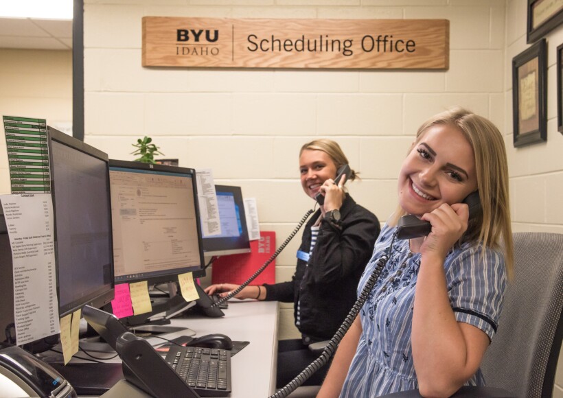 Two female students employees answer telephones in the Scheduling Office at BYU-Idaho.