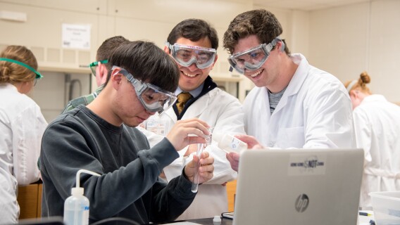 Students in protective gear help each other mix solutions in the chemistry lab.