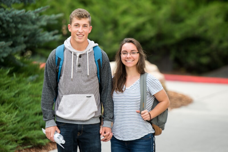 A male student wears a sweatshirt with a hood and and holds the hand of a female student wearing a striped shirt with sleeves. They smile for the picture.