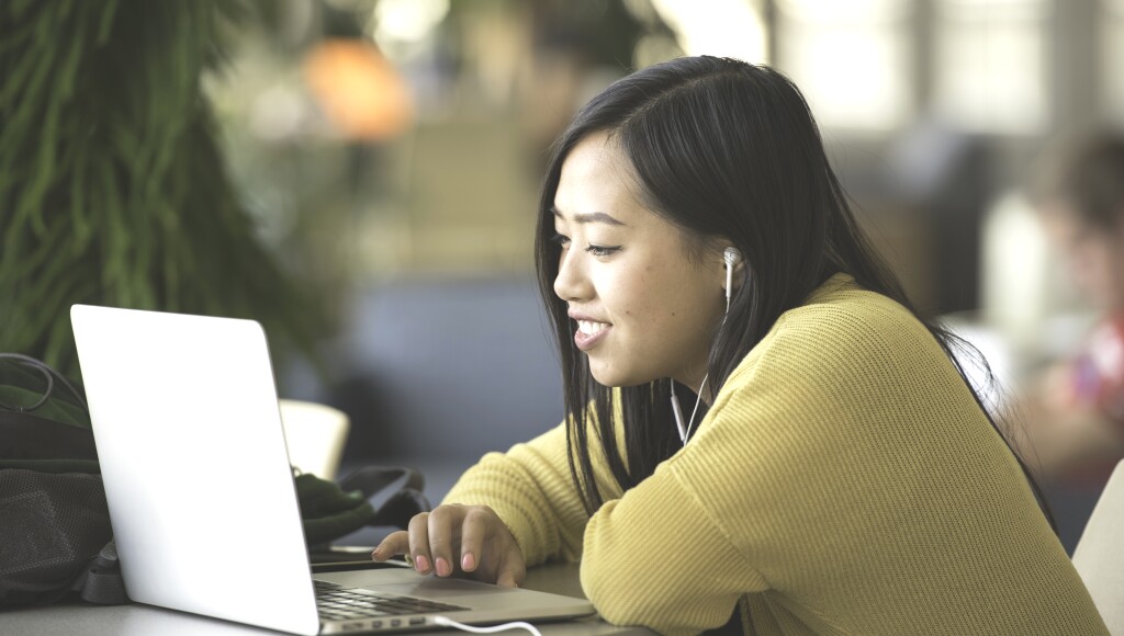 A female student with earbuds looks at a laptop and smiles.