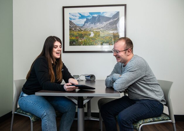 A male and female student sitting across from each other at a table, looking at an ipad.