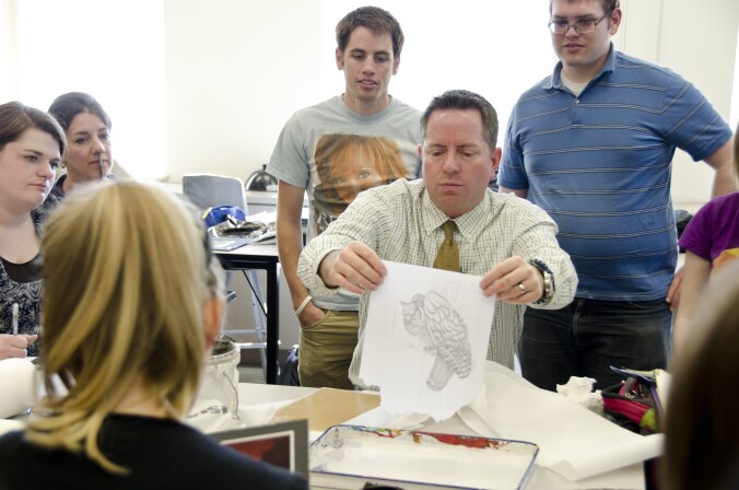 Students gather around their professor as he shows them a technique for illustration.