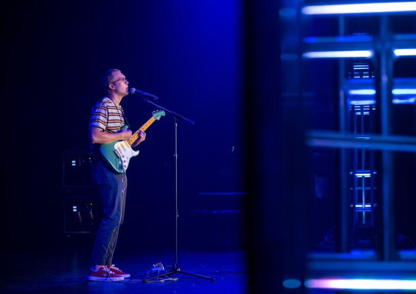 A BYU-Idaho student standing on a blue lit stage, holding a guitar an singing into a microphone.