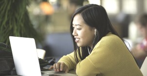 A female student with earbuds looks at a laptop and smiles.