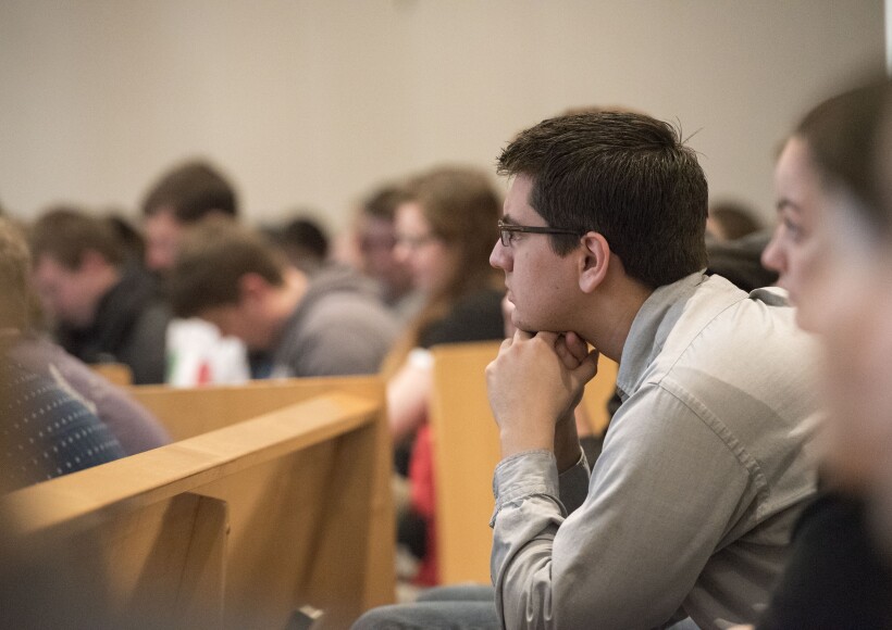 a man leaning forward with his elbow on his knees and his fists under his chin, sitting in a pew, listening to a speaker