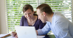 A married couple smile as they use a computer and internet at a kitchen table.