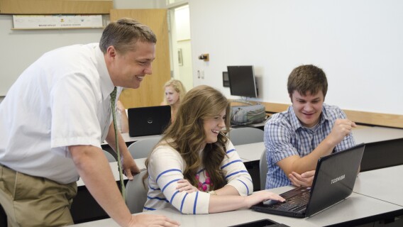 Two students smile while viewing a computer with a faculty member in a social studies education history class