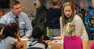 A student teaches a small group of children at a table with a supervisor looking on.