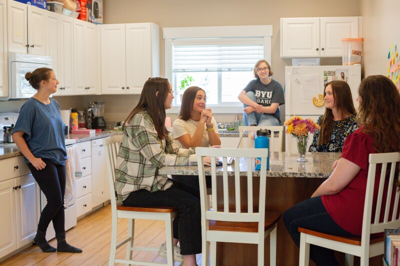 Female roommates all gathered together in the kitchen talking to one another.