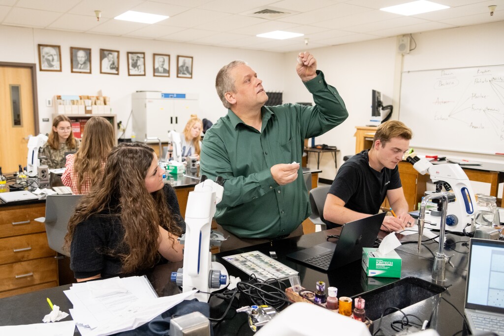 A professor examines something in a classroom with students working with microscopes.