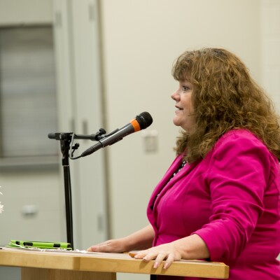 A women standing at a podium speaking to the women at the University Women's Association Luncheon.