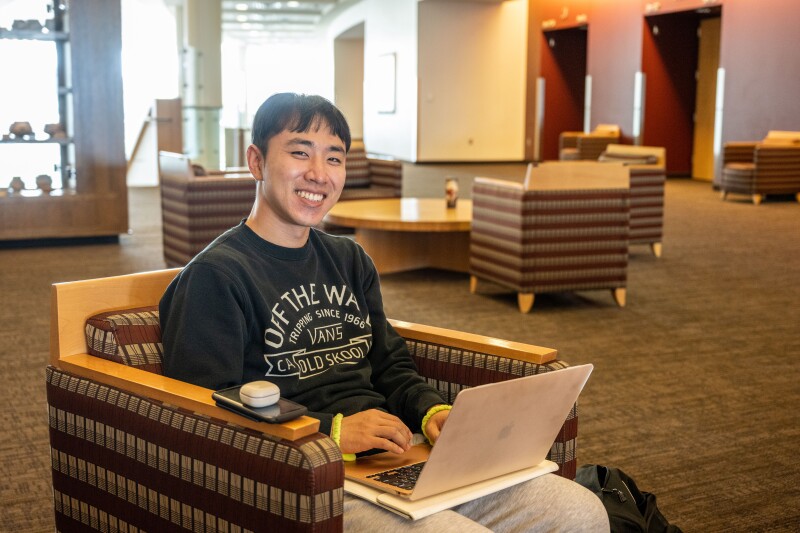 A BYU-Idaho student sitting in a chair with his laptop on his lap, smiling at the camera.