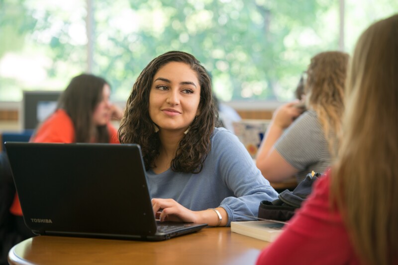 A female student working on a laptop at a table smiles while listening intently to a friend.