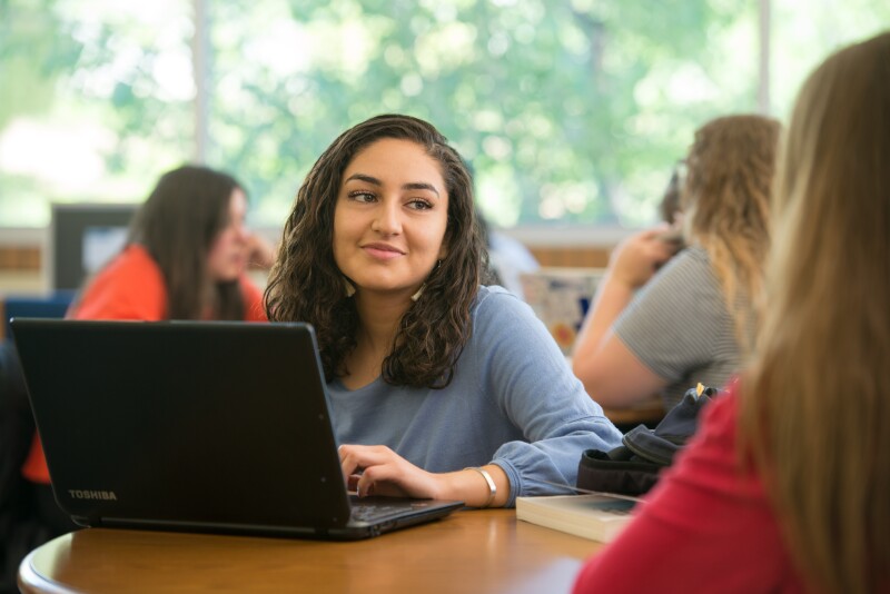 A female student working on a laptop at a table smiles while listening intently to a friend. 