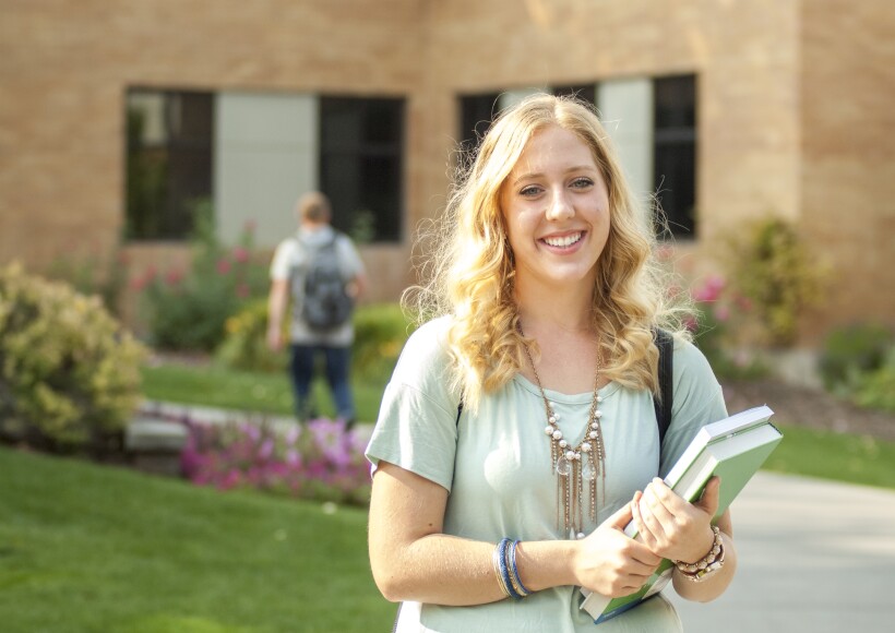 A female student in the Inter-University Relations program smiles outside a campus building carrying books for her classes.