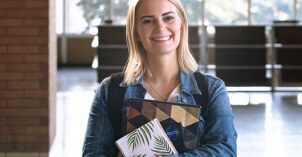 A Communication student stands in the Hart building with her notes and smiles.
