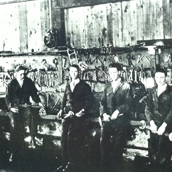 A vintage photograph from 1915 featuring five male students dressed in coveralls and hold various tools in a shop class.
