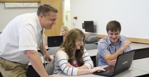 Two students smile while viewing a computer with a faculty member in a social studies education history class