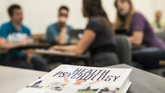 Image of a Health Psychology class. The textbook, Health Psychology, is in focus and students talk in the background.