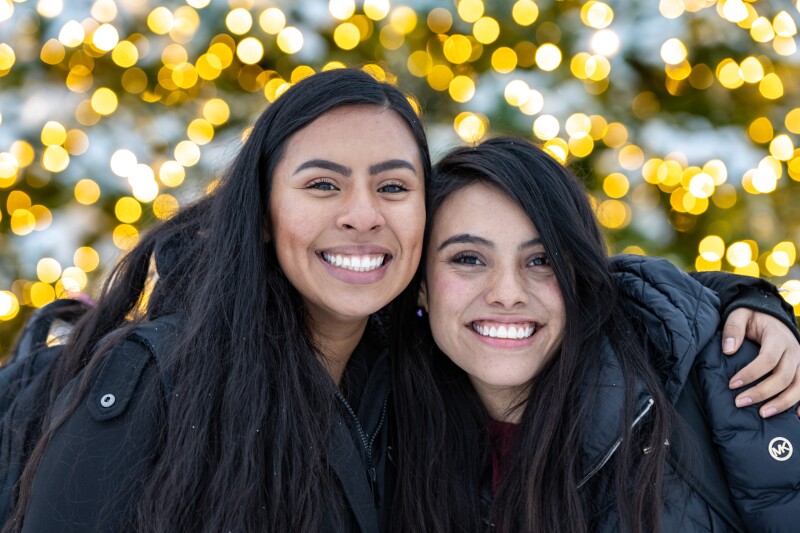 Two international students at BYU-I pose for a picture, twinkling Christmas lights in the background.